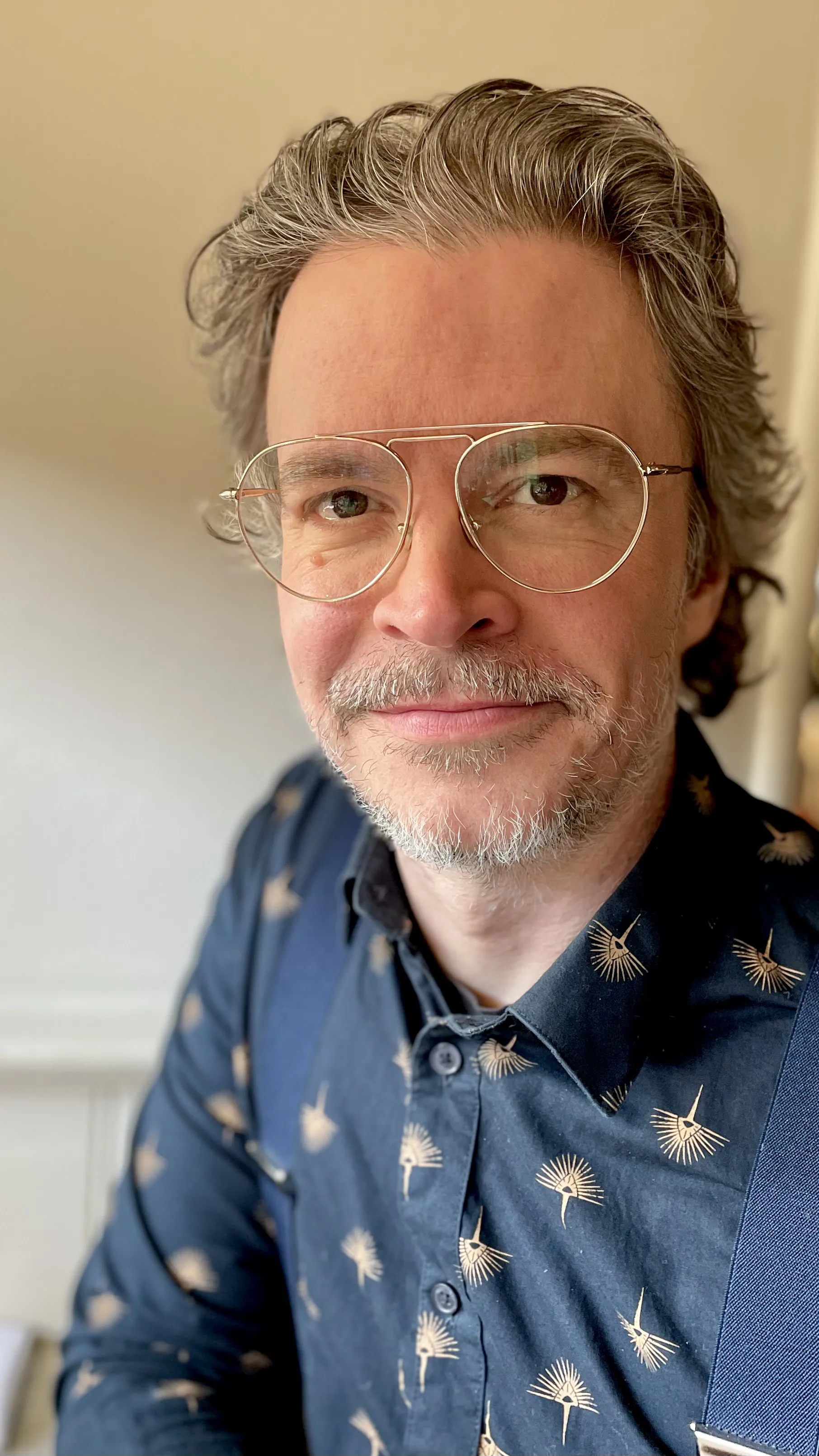 Närbild på en man med grånande, vågigt hår och mustasch. Han bär guldfärgade glasögon, en mörkblå skjorta med guldfärgade solfjäderformade figurer, och blå hängslen. Han ler lätt mot kameran.
